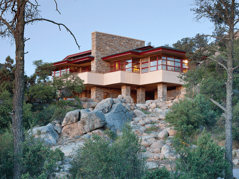 A multi-level stone-and-stucco house with red window frames, lit at dusk atop a hillside of granite boulders and pines.