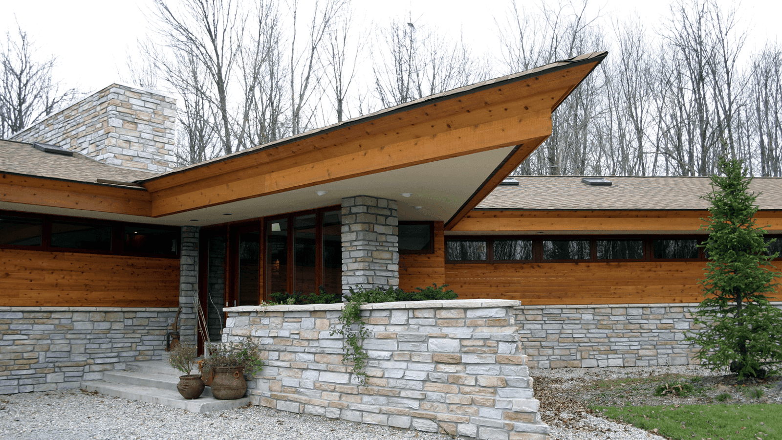 A single-story house with cedar siding and a stone base under a low cantilevered roof, set among bare trees.