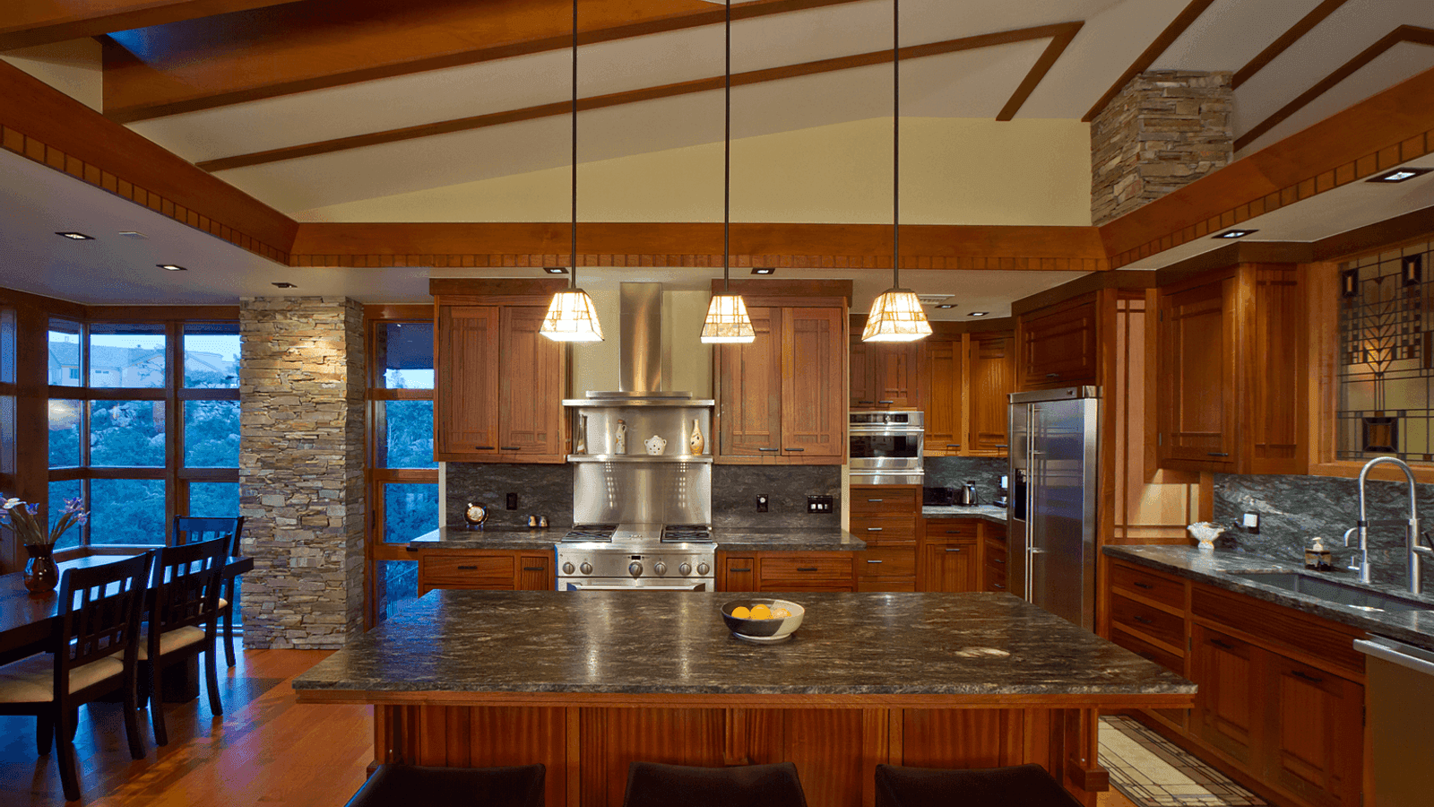 A warm-wood kitchen with exposed ceiling beams, three pendant lights, a stone column, and a large dark-granite island.